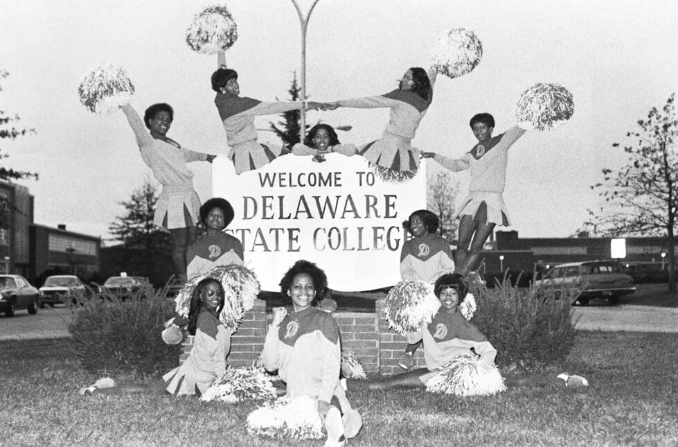 A 1973 photo of cheerleaders posing at the Delaware State College (now DSU) main entrance sign.